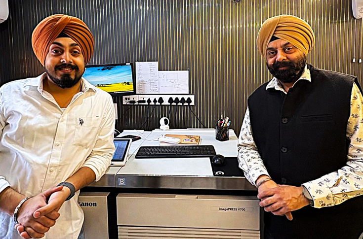 Family-run printing press with father and son operating modern Canon iPR V700 digital production machine alongside traditional and offset printing equipment, representing generational evolution in printing technology